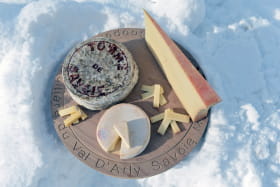 Fromages vus de haut (beaufort, tomme, reblochon) sur un plateau posé sur la neige.