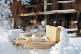 Fromages (beaufort, tomme, reblochon) sur un plateau posé sur la neige. En fond, un chalet traditionnel.