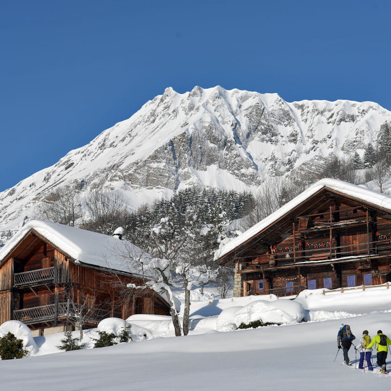 Trois personnes en raquettes marchent vers des chalets traditionnels sous la neige. En fond, la montagne enneigée se découpe sur le ciel bleu.